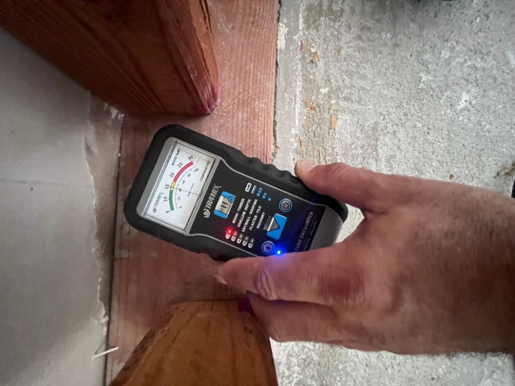 A hand holding a meter at the edge of a hardwood floor where it meets concrete in the East Bay