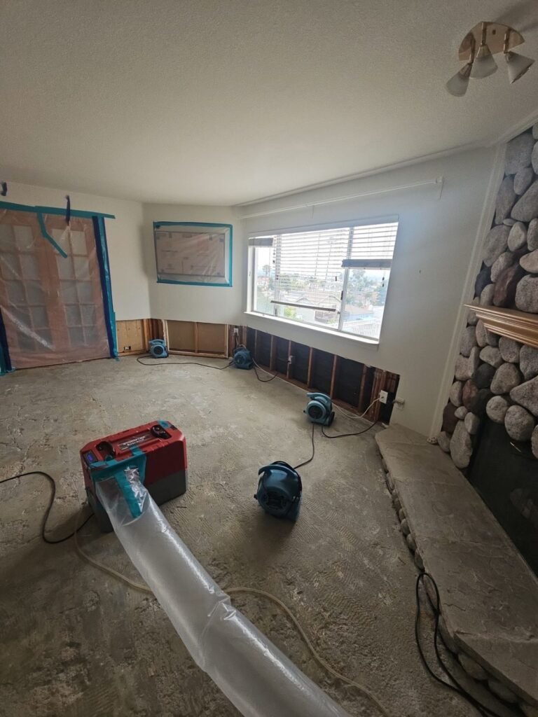 A room with the carpet pulled up to bare concrete, a red dehumidifier and ducting running to the window in the East Bay